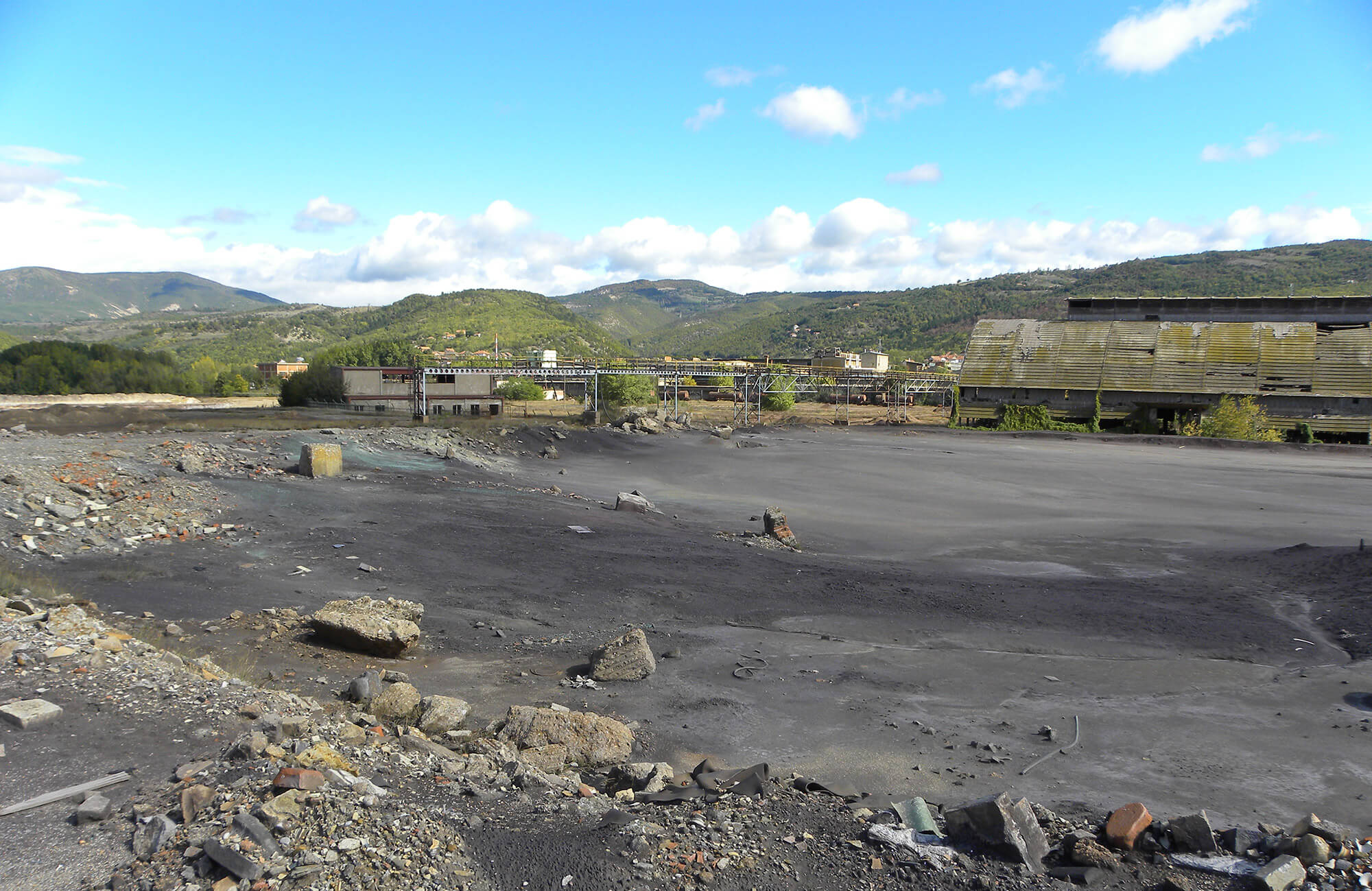Abandoned industrial site with dark, contaminated ground and scattered debris; former factory buildings and conveyor structures in the background, surrounded by hilly landscape under a partly cloudy sky.